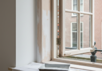 Een geopend houten raam met geschuurde kozijnonderdelen, een binnenmuur en schildermateriaal op een afdekdoek.